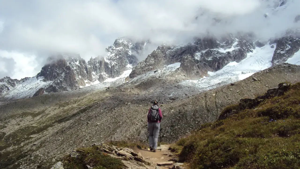 Osoba z plecakiem i kijkami idzie ścieżką w górach. W tle majestatyczne szczyty Alp, częściowo pokryte śniegiem i chmurami. Wspaniały widok na trekking w alpach.