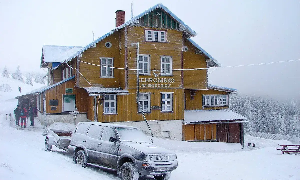 Schronisko na Śnieżniku w zimowej scenerii. Samochód z przyczepą i turyści przy wejściu. Masyw Śnieżnika i jego szlaki w śniegu.