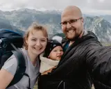 Rodzina na tle górskich szczytów. Uśmiechnięci rodzice z dzieckiem w nosidełku podziwiają czerwone wierchy.