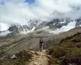 Osoba z plecakiem i kijkami idzie ścieżką w górach. W tle majestatyczne szczyty Alp, częściowo pokryte śniegiem i chmurami. Wspaniały widok na trekking w alpach.