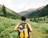 Kobieta z plecakiem wyrusza na trekking przez kwiecistą dolinę, podziwiając górskie krajobrazy.