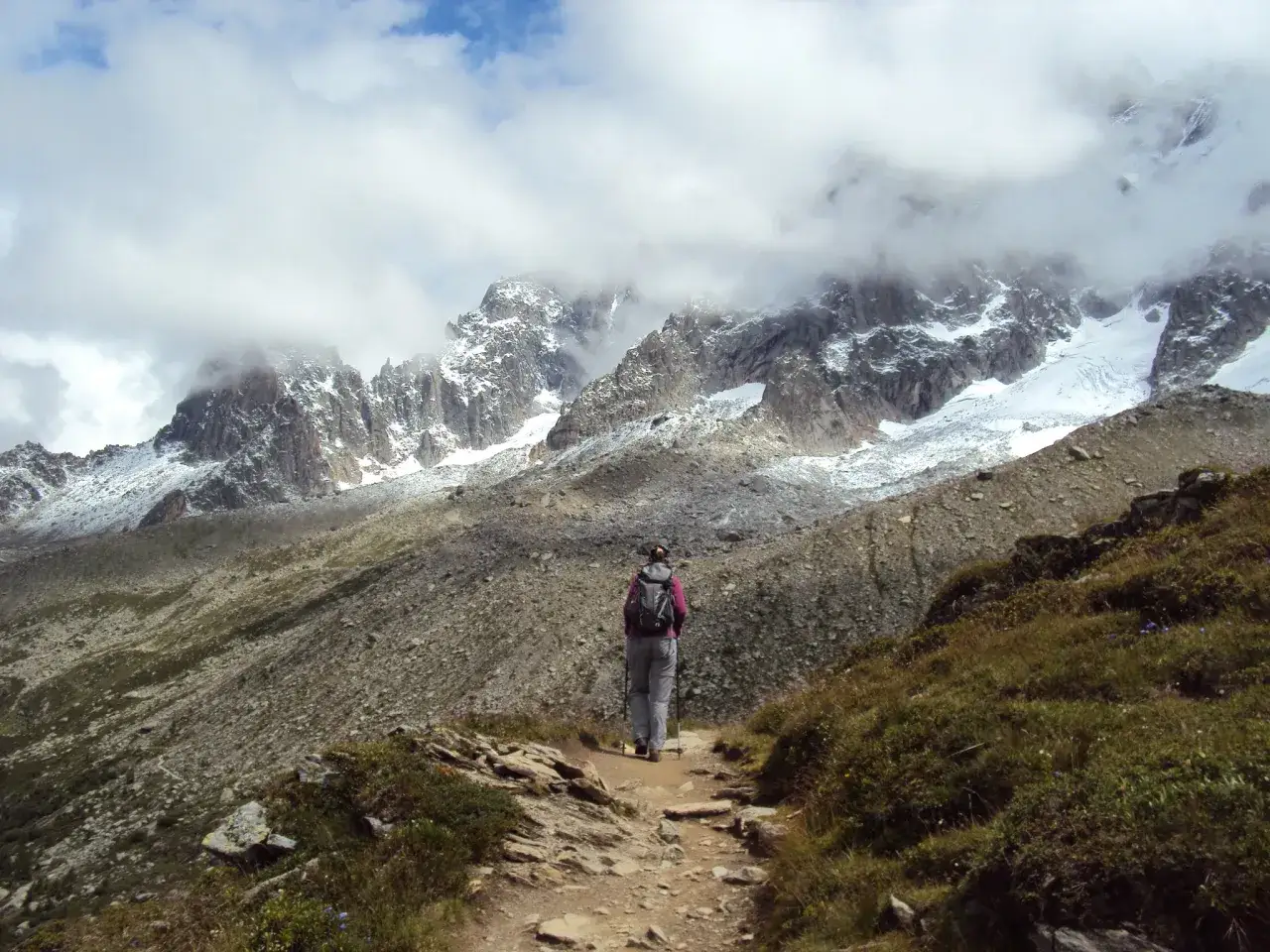 Osoba z plecakiem i kijkami idzie ścieżką w górach. W tle majestatyczne szczyty Alp, częściowo pokryte śniegiem i chmurami. To piękny dzień na trekking w alpach.
