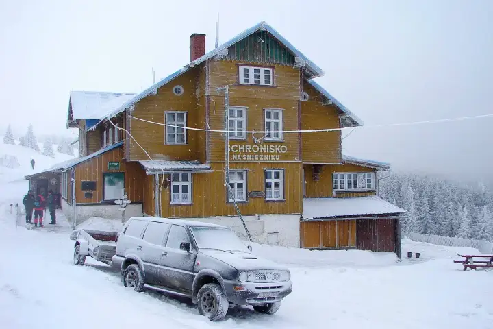 Schronisko na Śnieżniku w zimowej scenerii. Samochód z przyczepą i turyści przy wejściu. Masyw Śnieżnika i jego szlaki w śniegu.